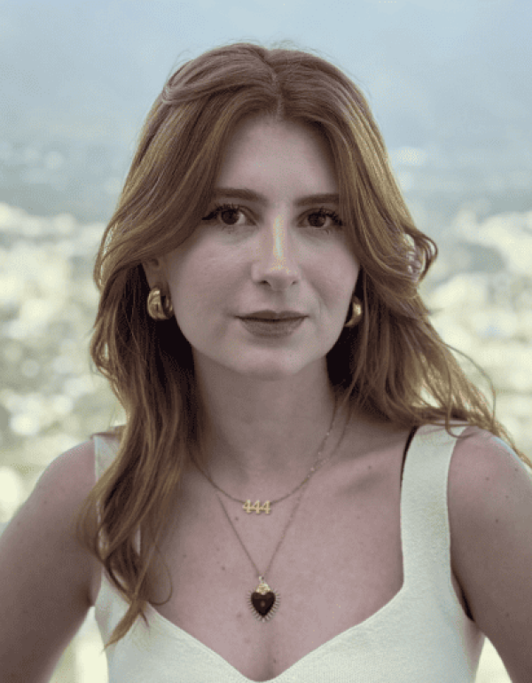 A young woman with red hair, wearing a white top, gold earrings, and layered necklaces, looks at the camera. A blurred cityscape and mountains are in the background.