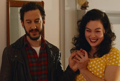 Mid-shot of a Caucasian man on the left with brown hair and beard dressed in a red and black check shirt and brown jacket as a smiling East and South East Asian woman stands to his right with curled hair and red lips wearing a yellow and white polka dot 50s style dress, holding his hand up towards her, while they both look slightly downwards towards the floor.