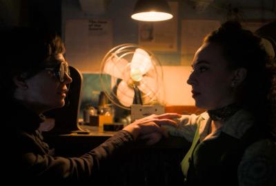 A woman dressed in black with opaque sunglasses holds hands with a film projectionist in a green vest. 