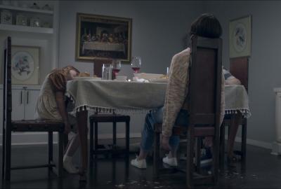 Taylor (Katie Parker) sits at the head of the table, her back to the camera, as the rest of the family surrounds her — all are eerily still in the dim light of the dining room. 