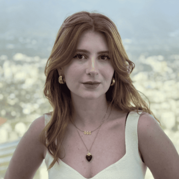 A young woman with red hair, wearing a white top, gold earrings, and layered necklaces, looks at the camera. A blurred cityscape and mountains are in the background.